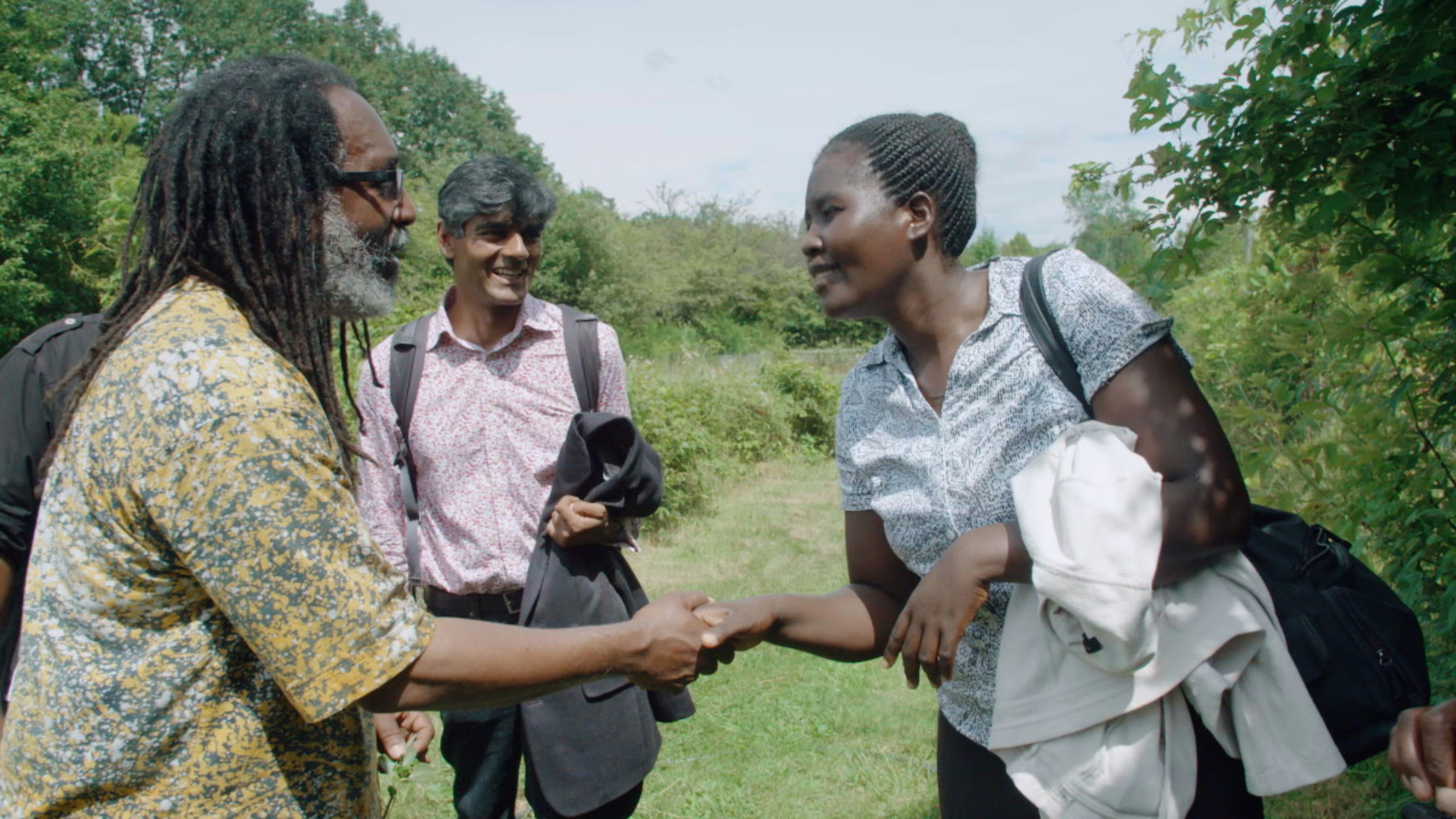 A man and woman shake hands in a lush African landscape while another man looks on smiling